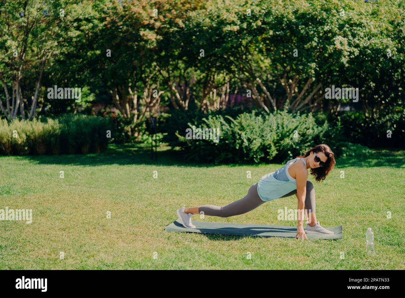 Concept de sport de plein air. Forme saine et énergique jeune femme fait des exercices de fitness sur le tapis de fitness habillé en vêtements actifs boissons l'eau douce porte des lunettes de soleil Banque D'Images