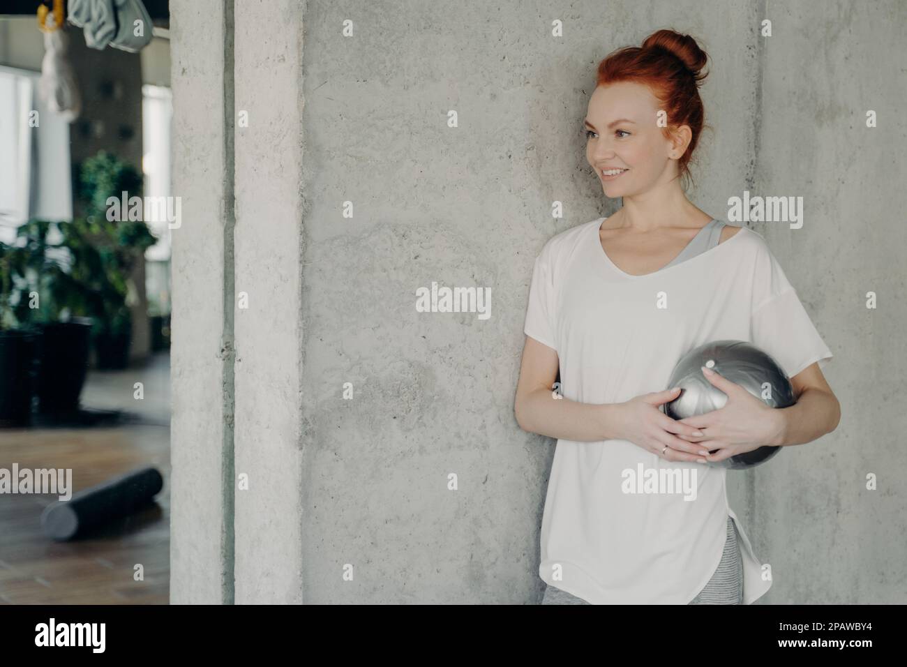 Femme aux cheveux rouges satisfaite en t-shirt de whire debout contre un mur de béton avec un petit fitball en main, appréciant le temps d'entraînement dans le studio de fitness, regardant Banque D'Images