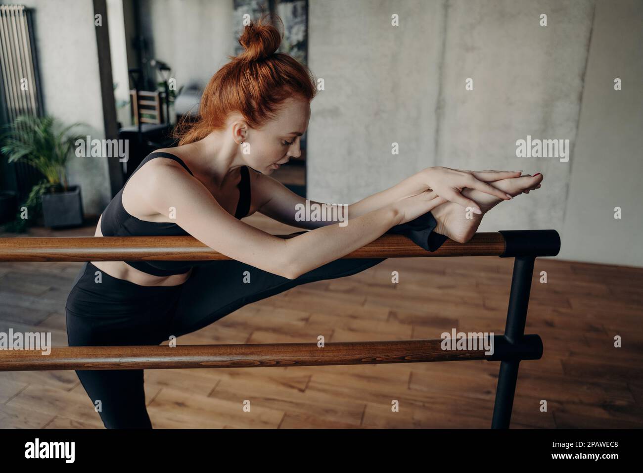 Jeune femme à cheveux rouges, mince ballerine ou proffesional en noir danseur de vêtements actifs s'exerçant dans le studio de fitness, avec les deux mains placées sur ankleman str Banque D'Images