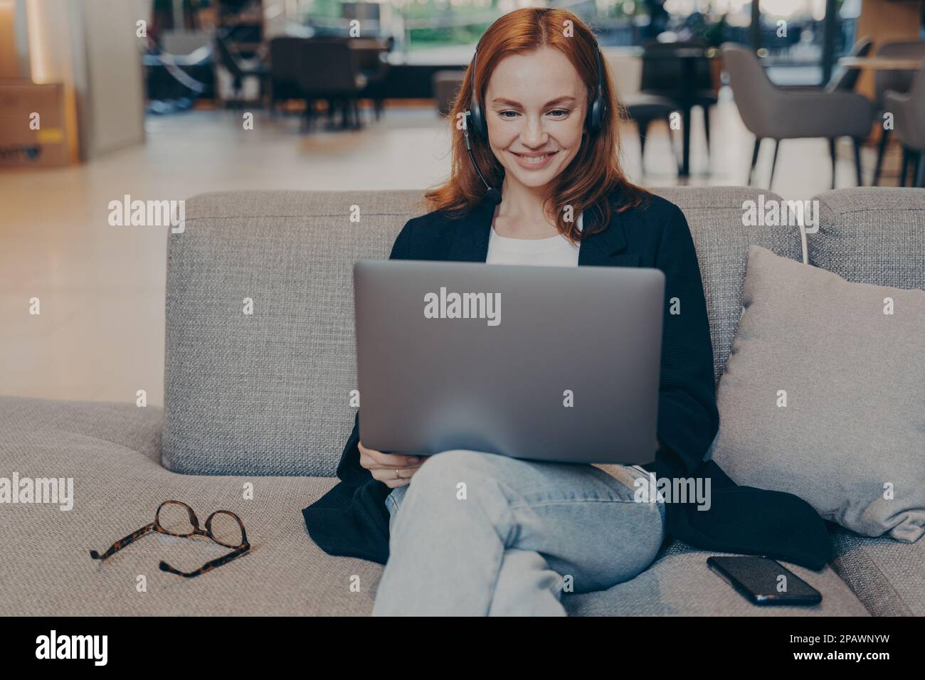 REDHEAD jeune femme communiquant avec les clients à l'aide d'un ordinateur portable et d'un casque sans fil, femme souriante parlant pendant un appel virtuel assis sur un canapé dans un café Banque D'Images