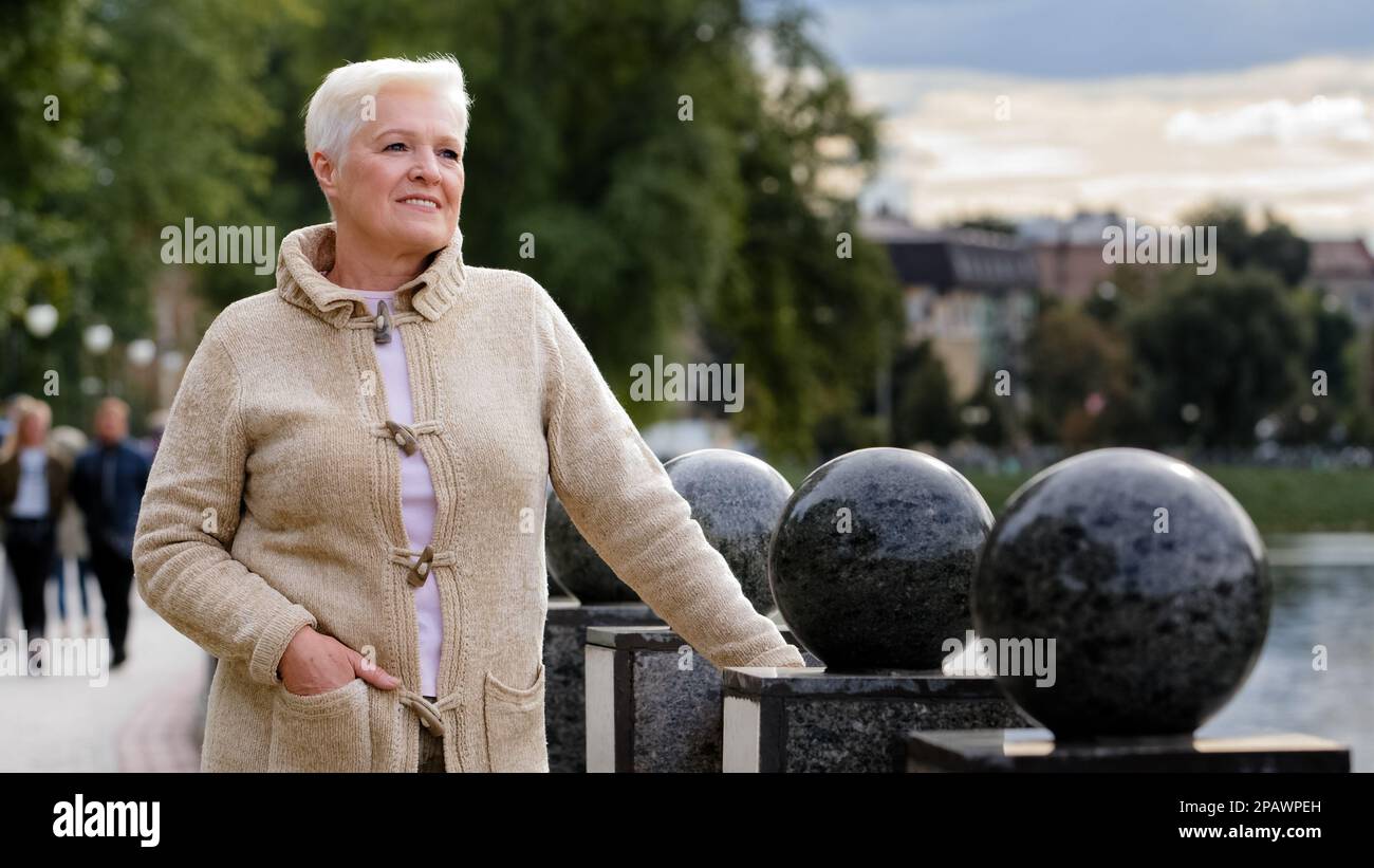 Femme aux cheveux gris retraite belle femme positive dans des vêtements chauds se détendre sur le remblai au bord de la rivière à l'extérieur par temps frais, en regardant loin sourire Banque D'Images