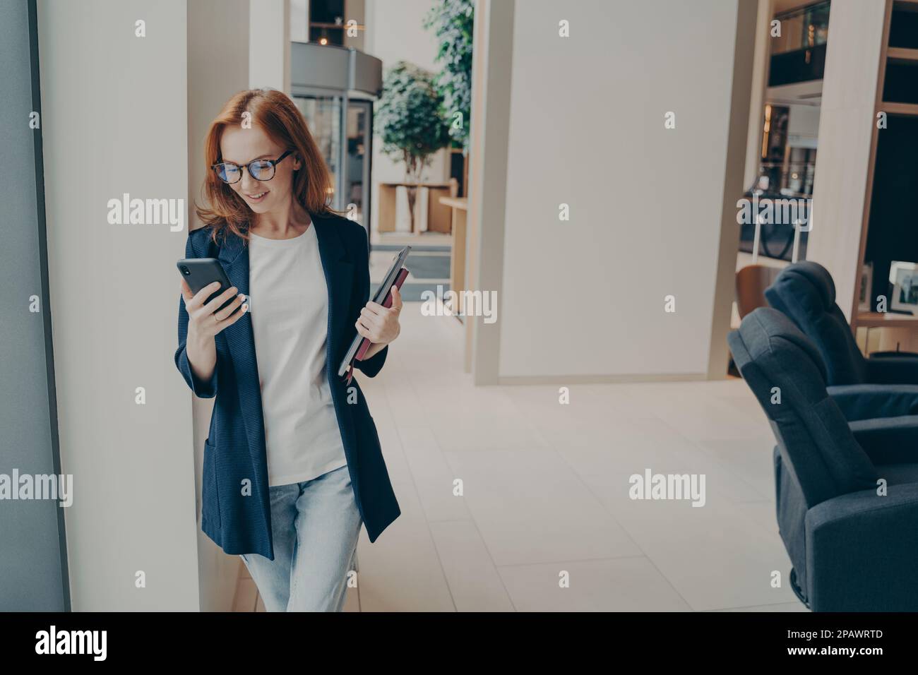 Souriante redhead femme consultant en affaires vêtue formellement tenant le téléphone mobile, lisant les nouvelles en ligne, debout au bureau pendant la pause-café ou après Banque D'Images
