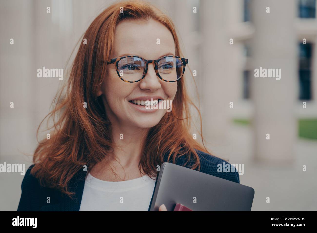 Portrait d'une femme élégante et heureuse avec un large sourire en regardant de côté en attendant son partenaire d'affaires à l'extérieur, en tenant un ordinateur portable moderne et en portant un spectateur Banque D'Images