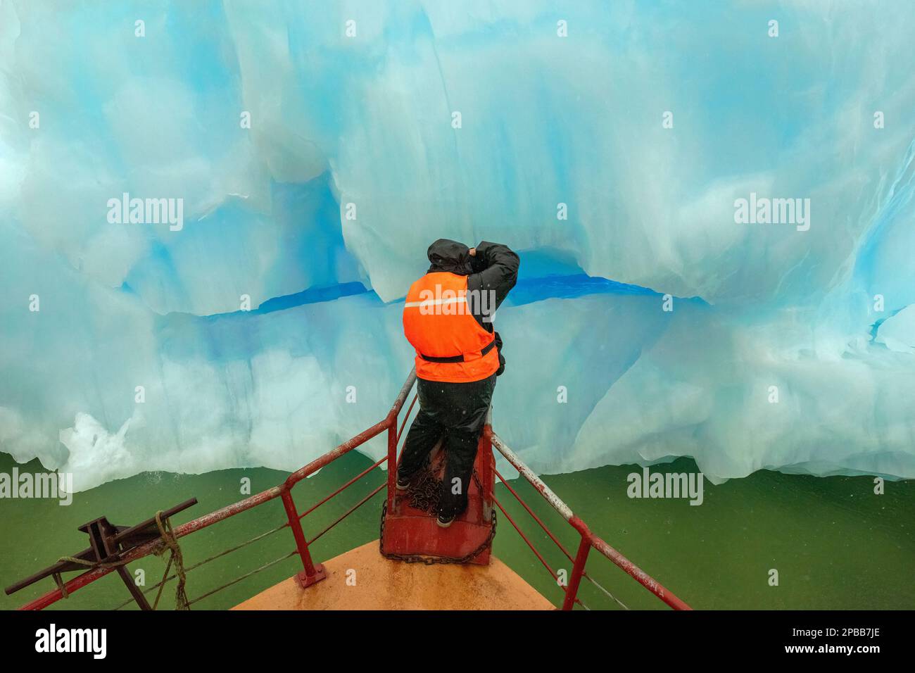 Photographe sur la proue du navire avec un iceberg bleu et de l'eau verte remplie de plancton sous la pluie, le glacier Jorge Montt, Patagonie Banque D'Images