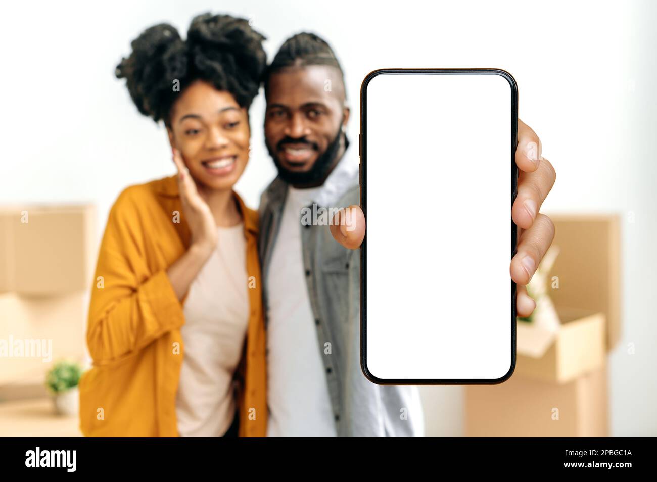 Couple afro-américain en mode flou artistique, montrez un smartphone avec un écran cache blanc vide tout en vous tenant dans le salon contre le fond de boîtes en carton avec des objets dans leur nouvelle maison Banque D'Images