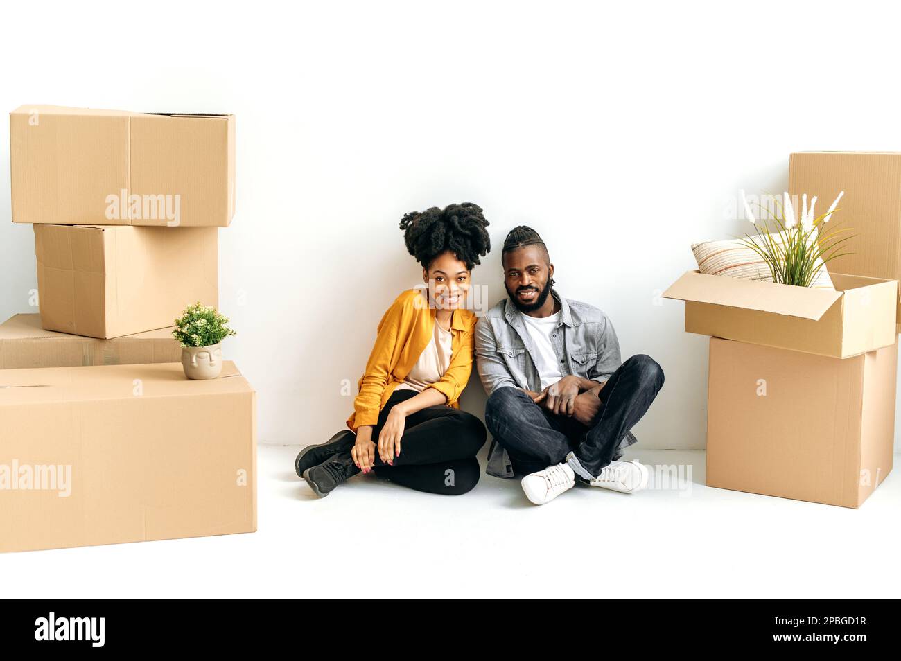 Joyeux couple africain américain marié, vêtu de vêtements décontractés, assis dans leur première maison sur le sol, il ya des boîtes à cardbox avec des choses autour, ils regardent la caméra, souriant, investi dans l'immobilier Banque D'Images