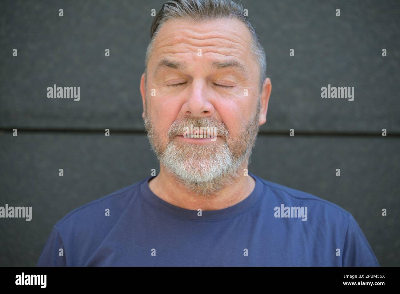 Homme barbu d'âge moyen posant avec les yeux fermés et les lèvres parted contre un mur sombre Banque D'Images