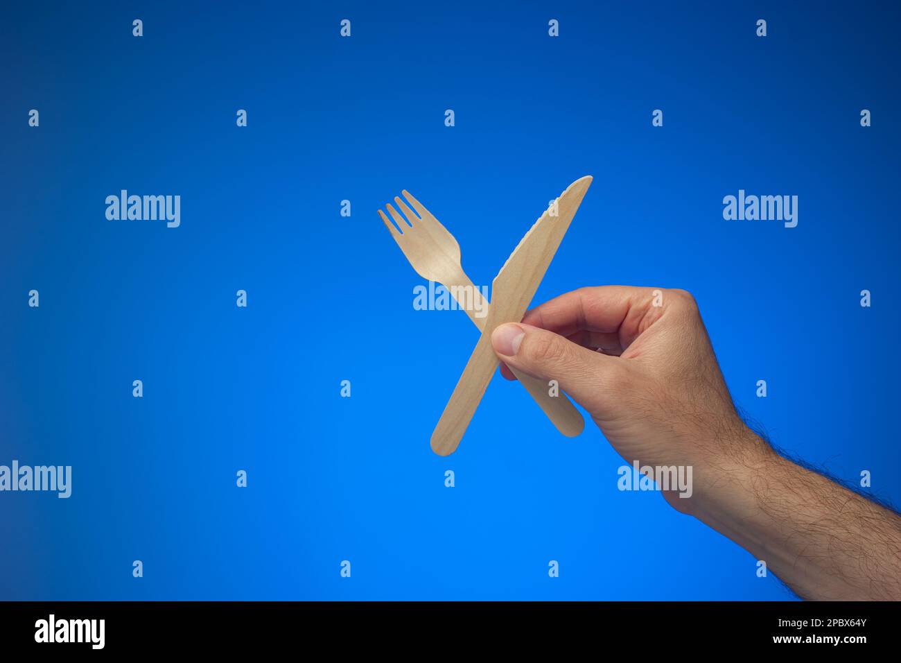 Couteau et fourchette en bois à usage unique, à usage unique et biodégradable, tenu en main par un homme caucasien. Gros plan en studio, isolé sur fond bleu. Banque D'Images