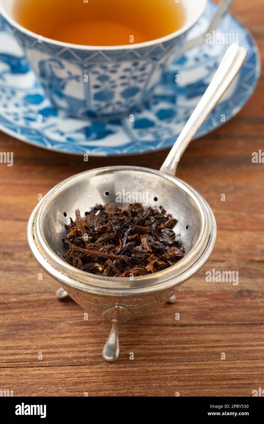Ancienne passoire en argent et support avec feuilles de thé mouillées devant une tasse de thé sur une table en bois Banque D'Images