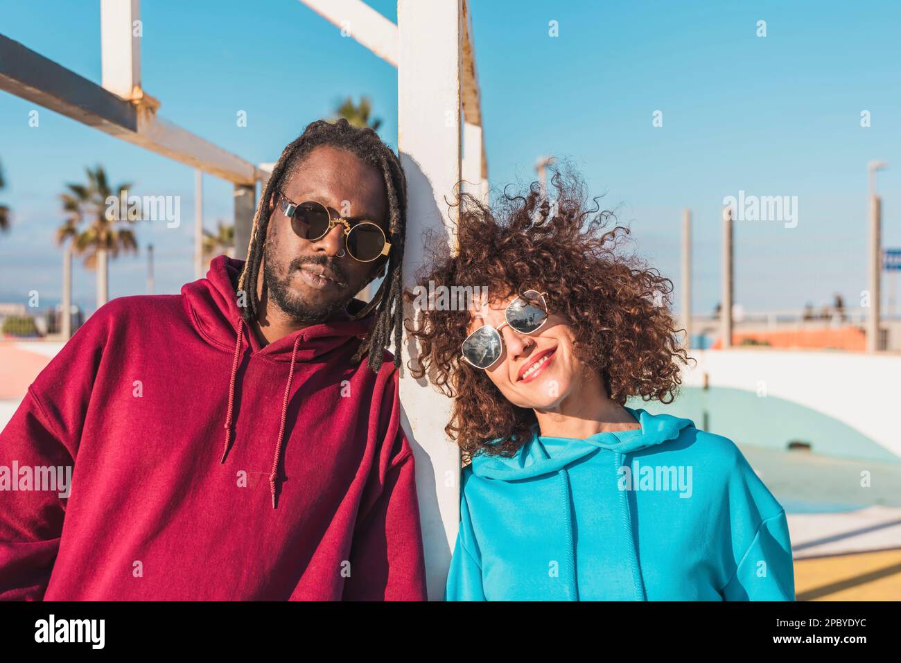 Heureux couple multiracial dans des sweats à capuche lumineux souriant et regardant l'appareil photo tout en se tenant dans la rue contre le ciel bleu sans nuages ensemble Banque D'Images