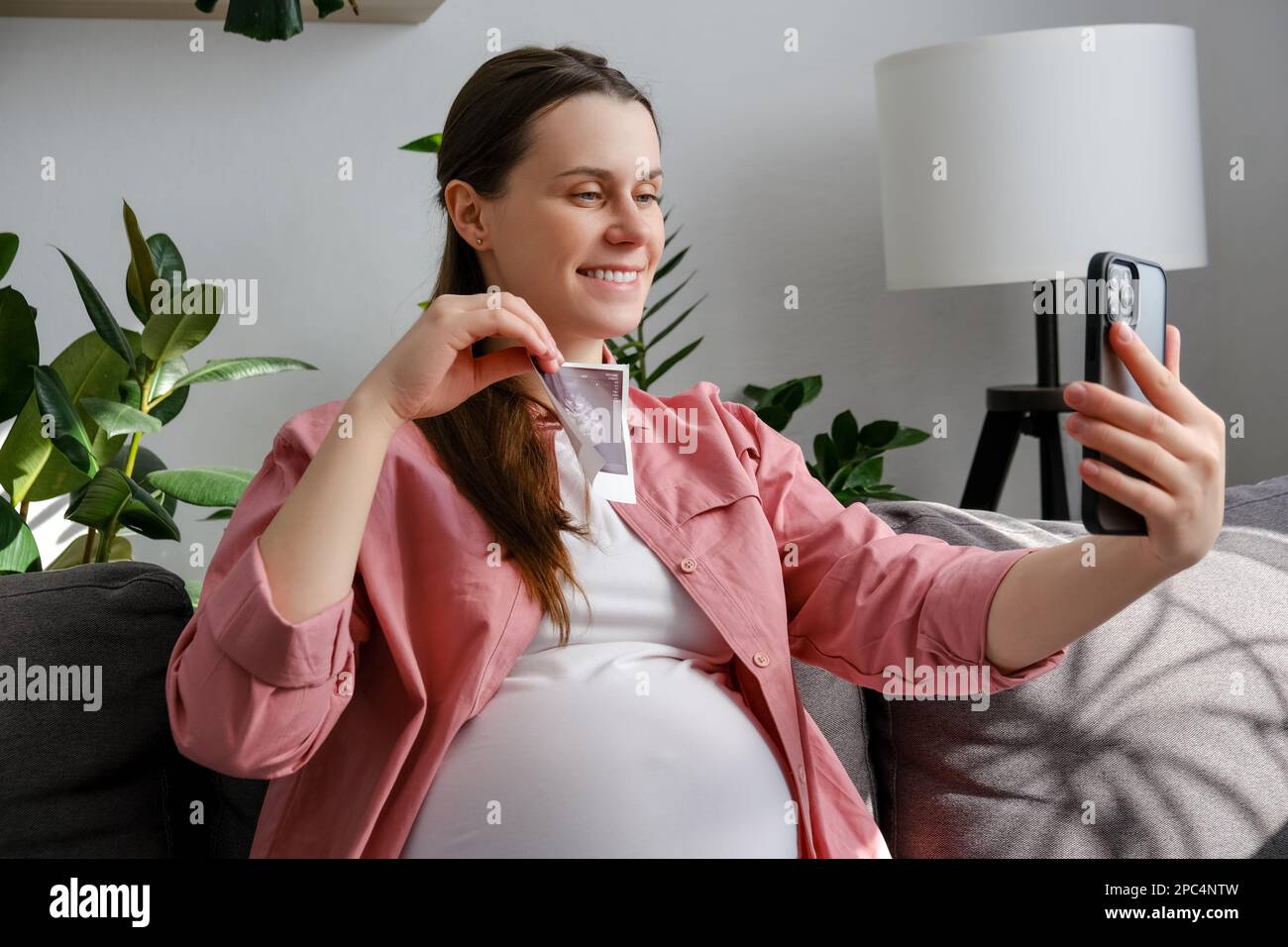 Gros plan d'une belle jeune femme enceinte souriante assise sur un canapé confortable tenant des images échographiques de bébé avec sa famille à l'aide d'un smartphone avec vidéo Banque D'Images