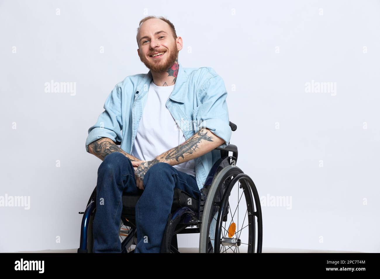 Un homme dans un fauteuil roulant et pointant un doigt, un espace de copie, avec des tatouages sur ses mains est assis sur un fond gris studio, concept de santé une personne avec Banque D'Images