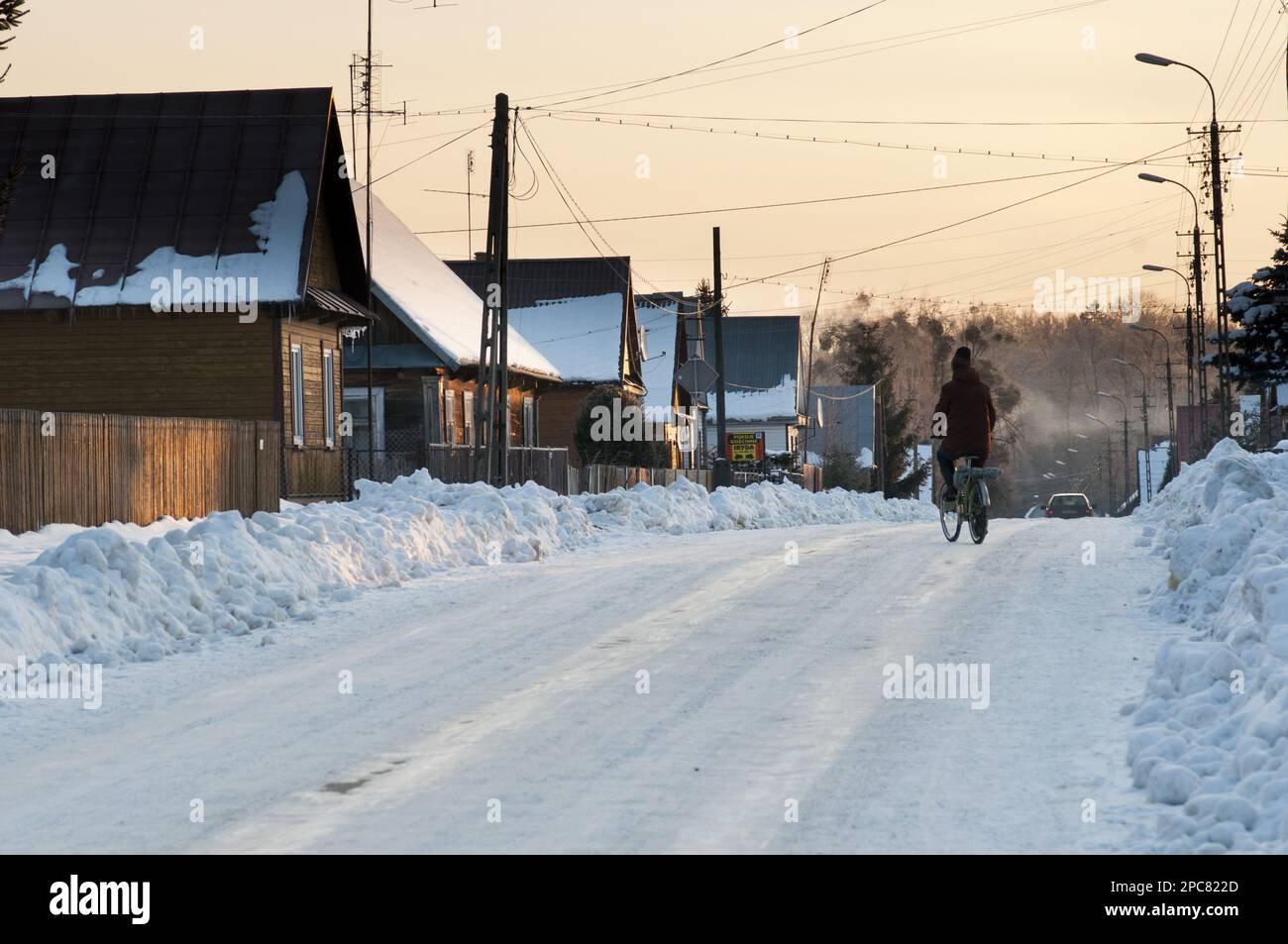 Cycliste sur route enneigée, maisons traditionnelles en bois et tas de neige défrichie, Bialowieza Village, Podlaskie Voivodeship, Pologne Banque D'Images