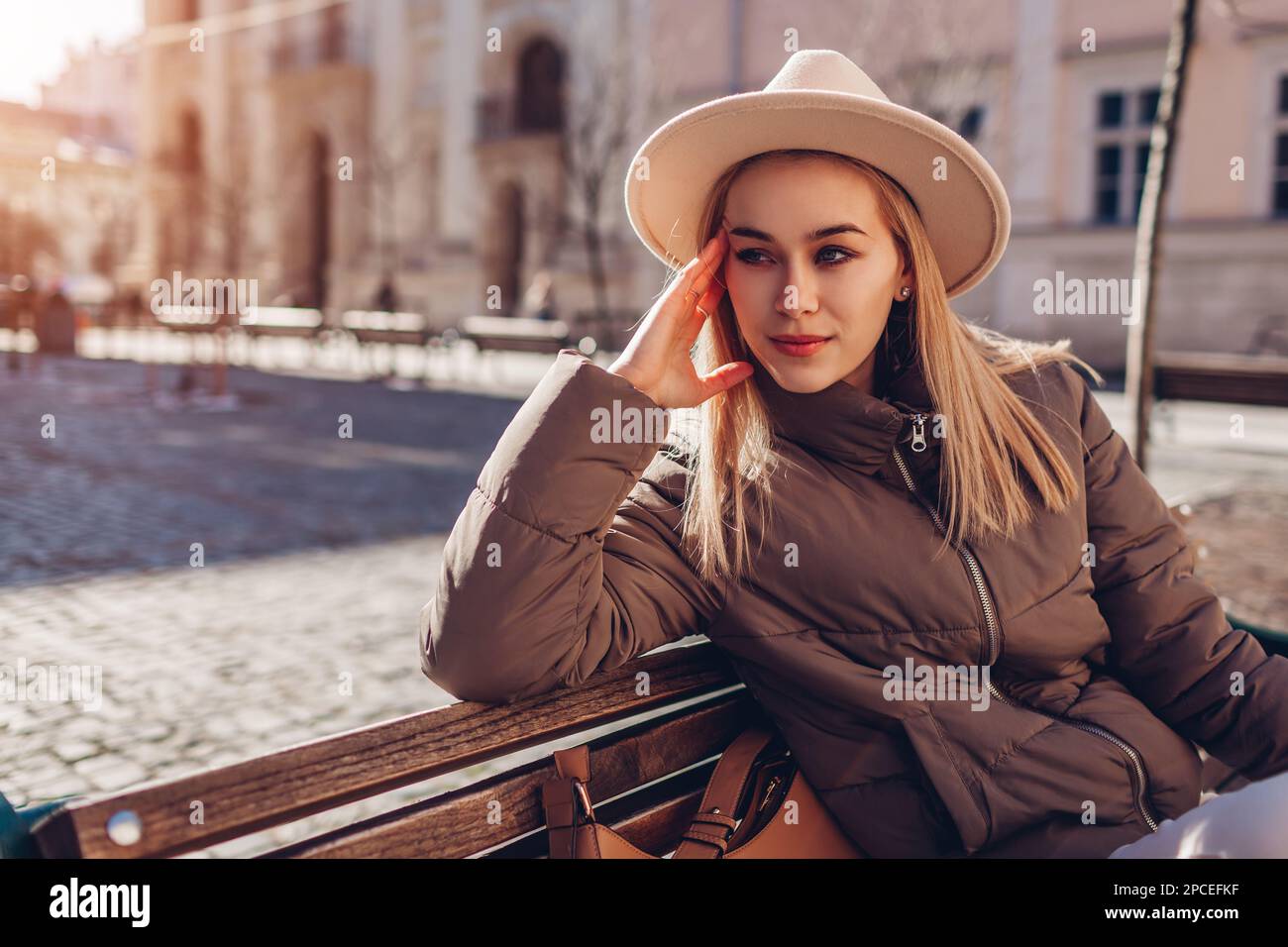 Jeune femme à la mode assise sur un banc dans la rue de la ville  européenne, portant un élégant chapeau et une veste tenant un sac à main.  Tenue de printemps. Beige