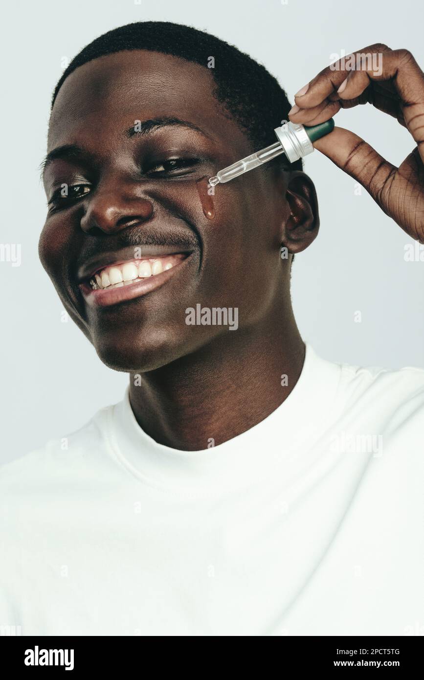 Portrait d'un heureux, jeune homme noir souriant tout en appliquant le sérum de visage avec un compte-gouttes de beauté. Homme prenant soin de sa peau à l'aide d'un produit hydratant Banque D'Images