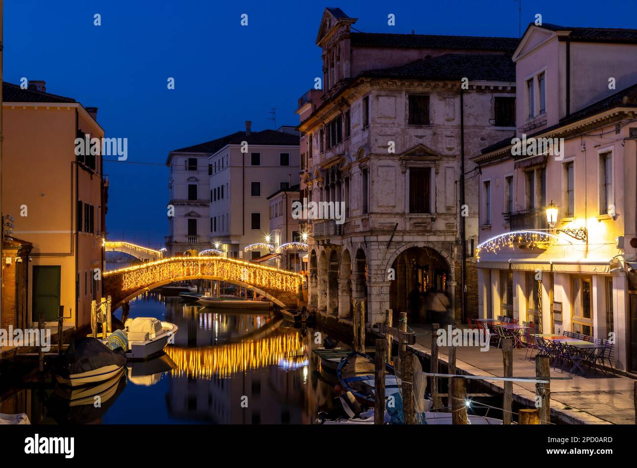 Chioggia paysage urbain dans la lagune de Venise avec étroit canal d'eau de Vena avec des bateaux colorés parmi les anciens bâtiments et pont de brique illuminé dans la soirée Banque D'Images