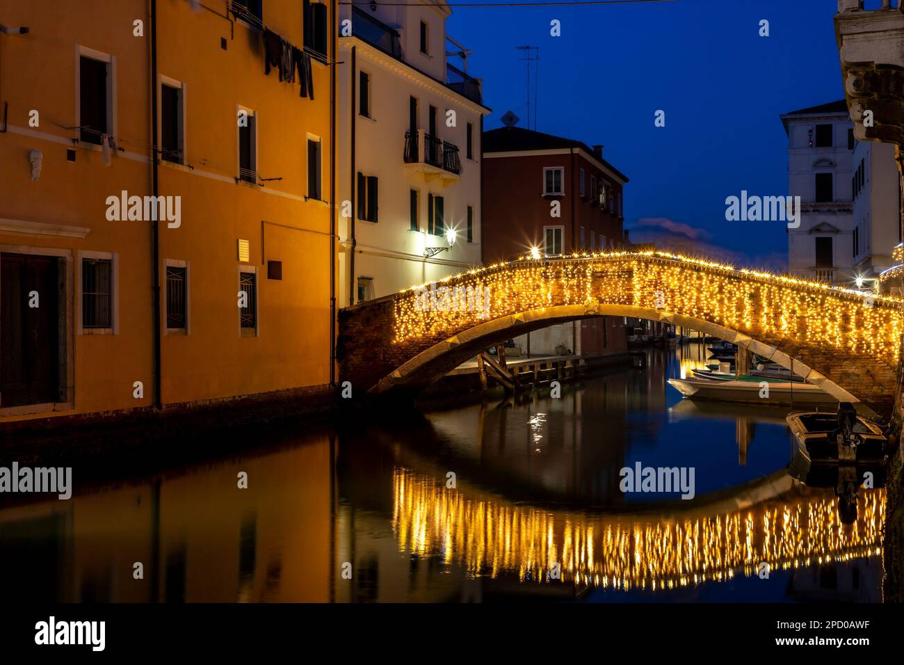 Chioggia paysage urbain dans la lagune de Venise avec étroit canal d'eau de Vena avec des bateaux colorés parmi les anciens bâtiments et pont de brique illuminé dans la soirée Banque D'Images