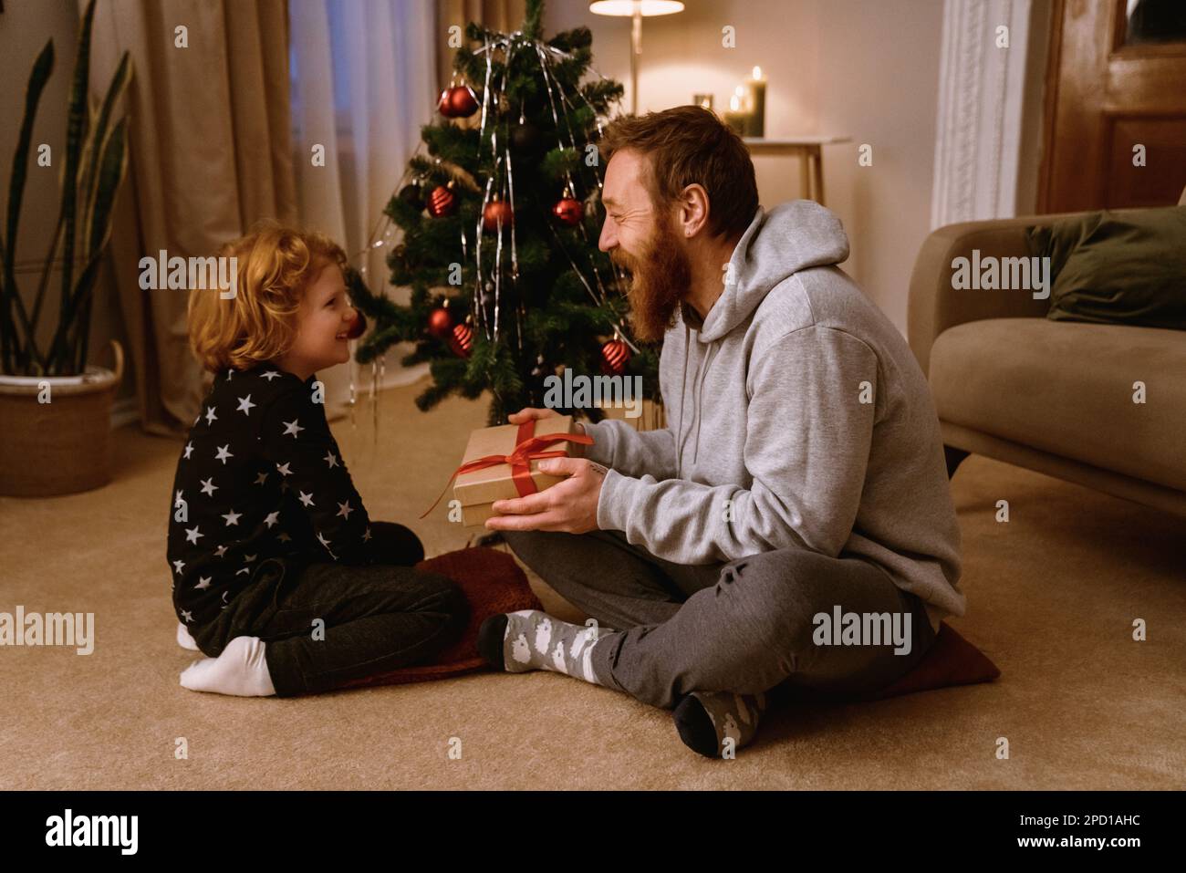 Mignon petit garçon souriant recevant le cadeau de son père tout en étant assis sur le sol dans le salon confortable avec arbre de Noël sur fond Banque D'Images