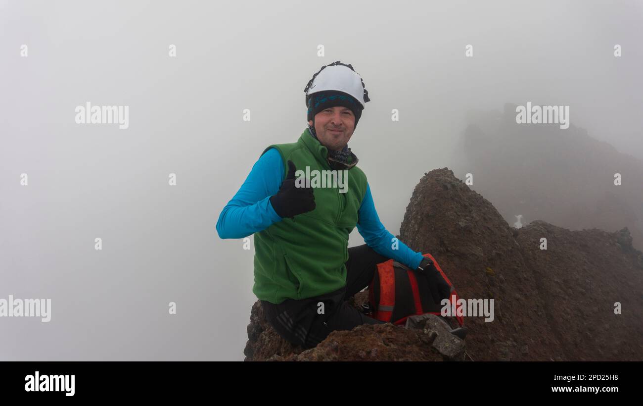 Jeune homme grimpeur assis au sommet du volcan Ruminahui portant un manteau vert et un casque par temps nuageux Banque D'Images
