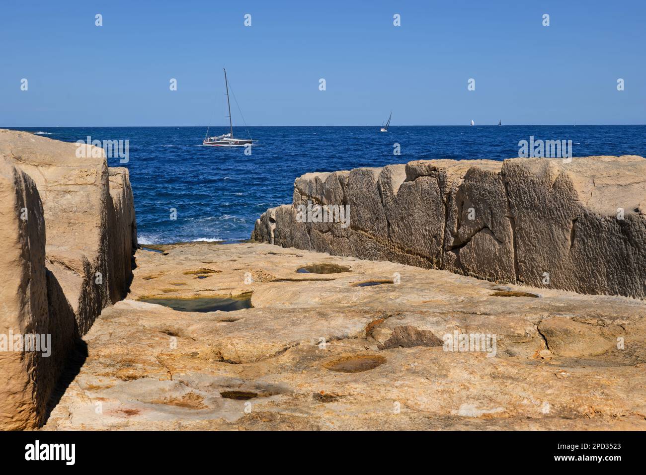 Rampe d'accès pour bateaux (lancement, déployeur de bateaux, cale) vers la mer avec bateau à voile à l'horizon, sculptée sur la rive rocheuse du bord de mer de la Valette à Malte. Banque D'Images