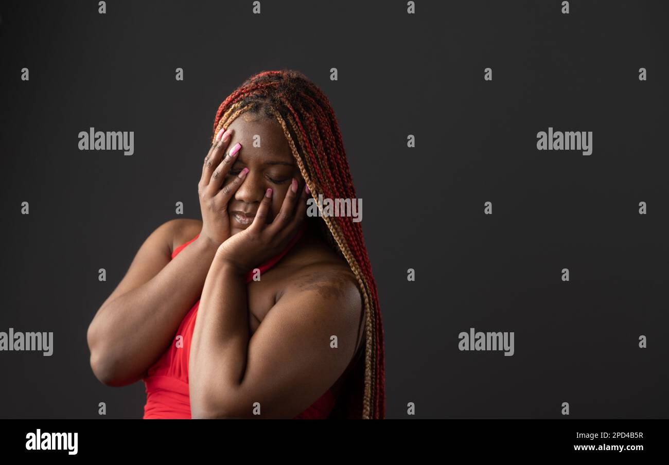 Une femme afro-américaine avec des tresses rouges couvrant son visage avec les mains sur un fond sombre Banque D'Images