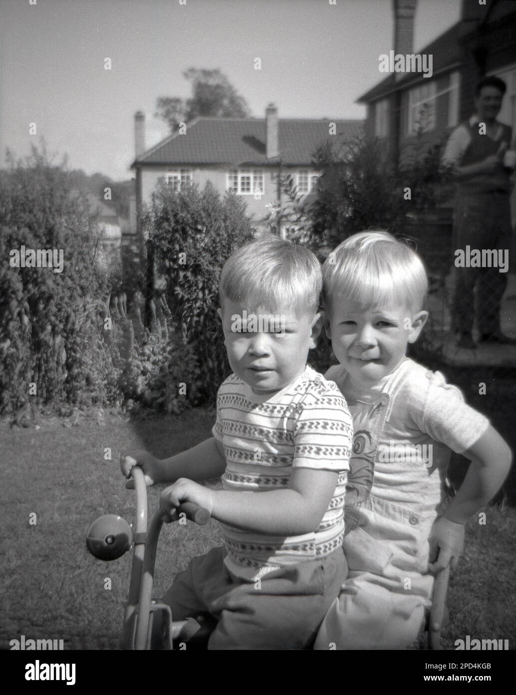 1964, historique, l'été, à l'extérieur dans un jardin, deux petits garçons blonds aux cheveux assis ensemble sur un tricycle d'enfant, Angleterre, Royaume-Uni. Banque D'Images