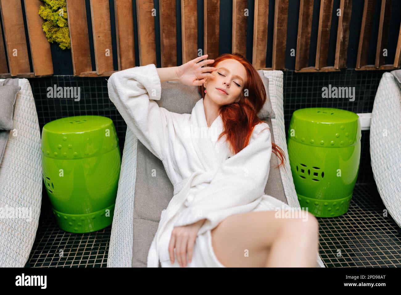 Portrait d'une jolie jeune femme à tête rouge portant un peignoir blanc sur le visage allongé avec des yeux fermés sur un transat après un soin spa au salon de beauté. Banque D'Images