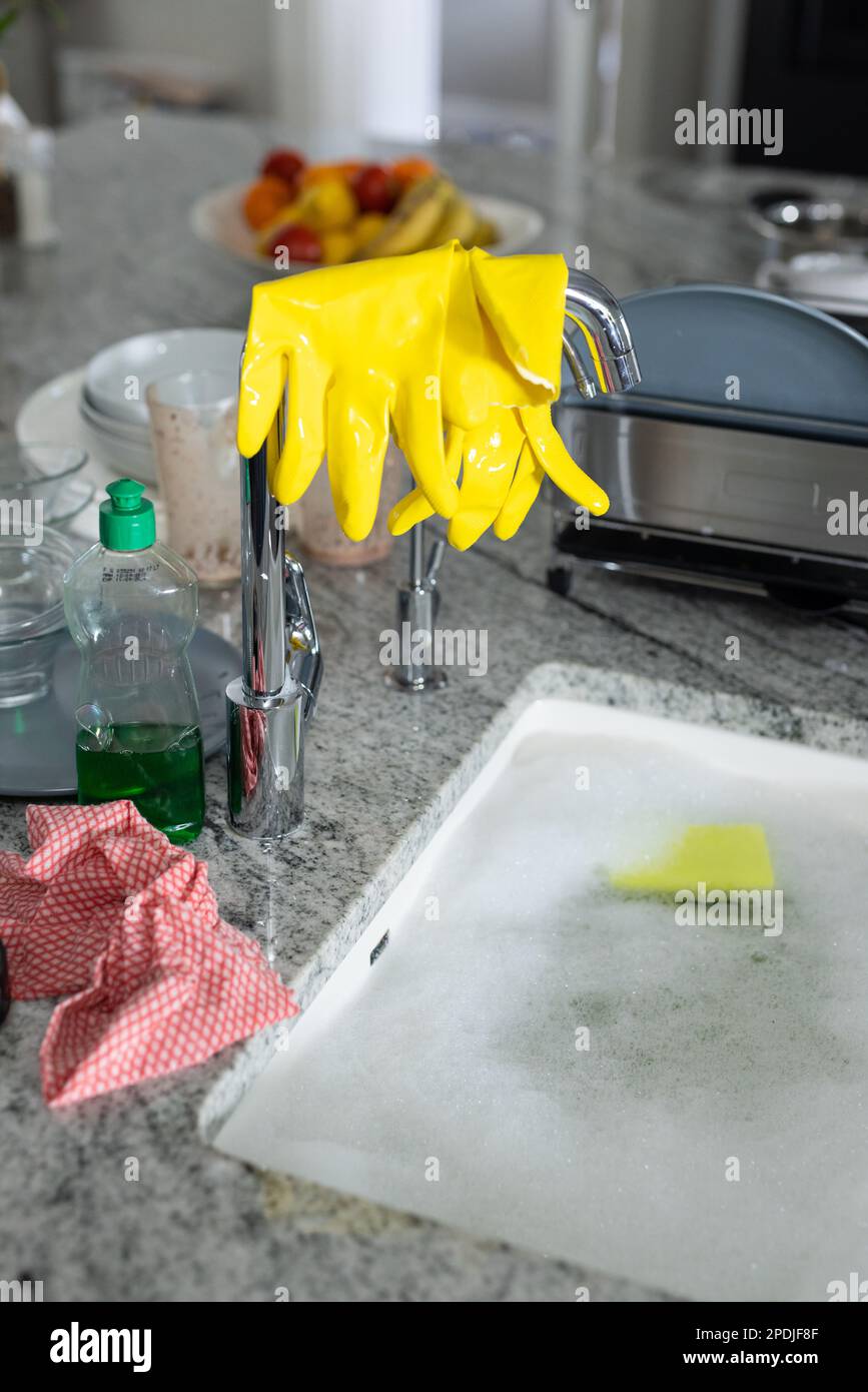 Distributeur de savon par gants de lavage jaunes sur le robinet et l'évier de cuisine rempli d'eau savonneuse Banque D'Images
