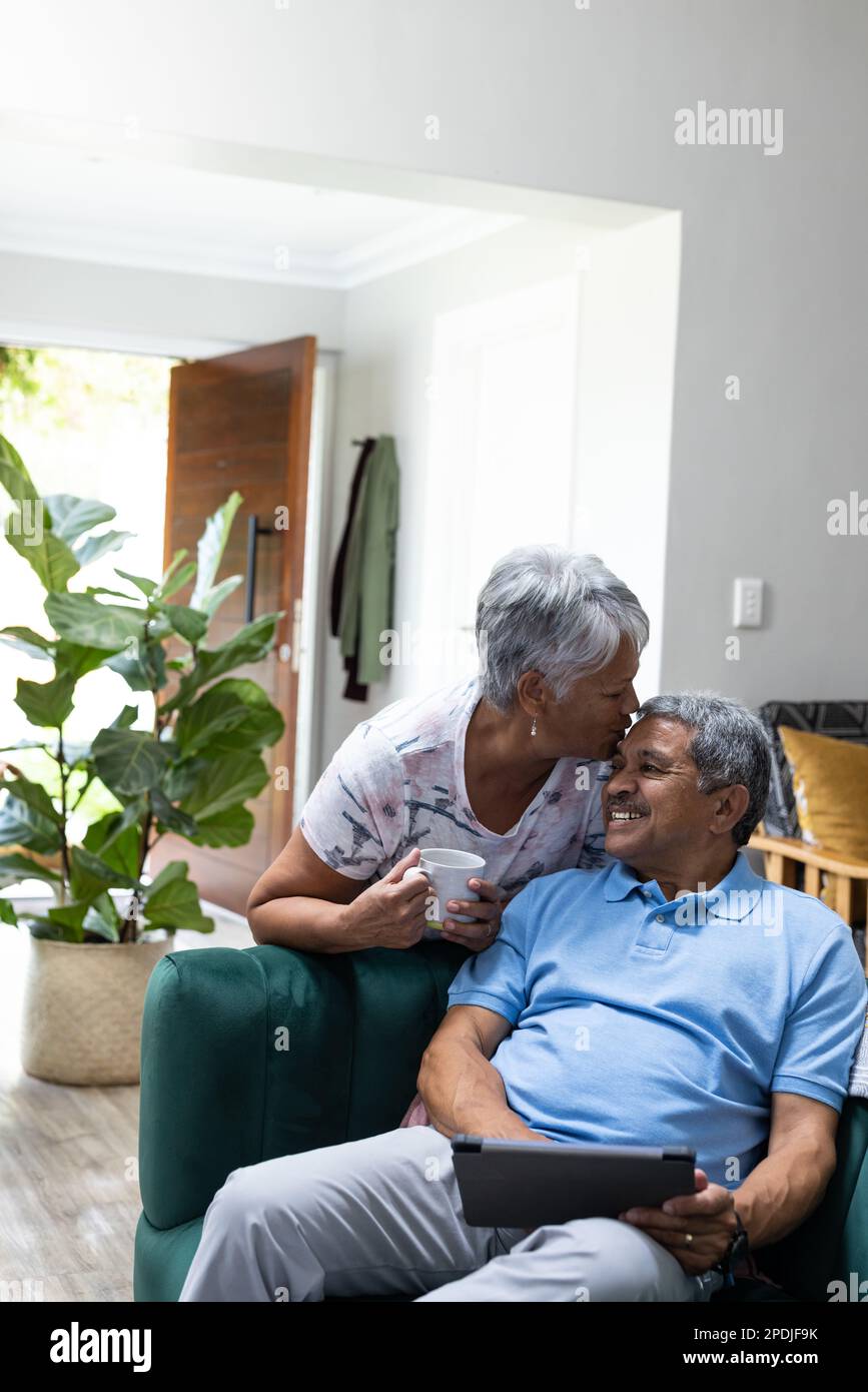Femme biraciale senior avec une tasse de café embrassant sur le front de son mari en utilisant une tablette numérique sur le canapé Banque D'Images