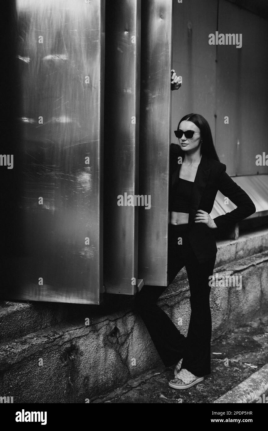 Portrait de mode de jeune femme élégante en plein air. Costume noir, lunettes de soleil, fond gris en métal. Banque D'Images