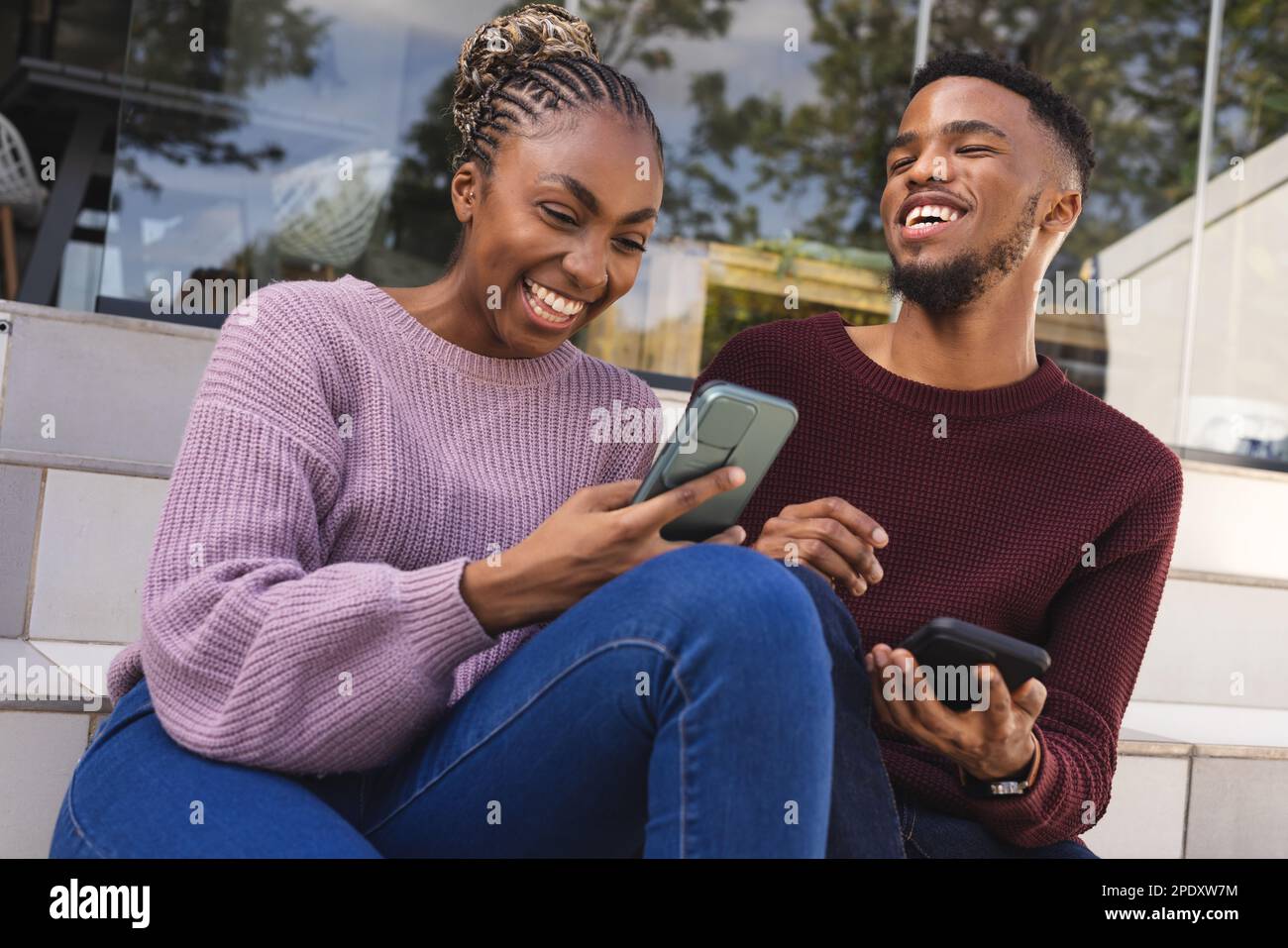 Heureux couple afro-américain assis sur les escaliers dans le jardin, en utilisant des smartphones Banque D'Images