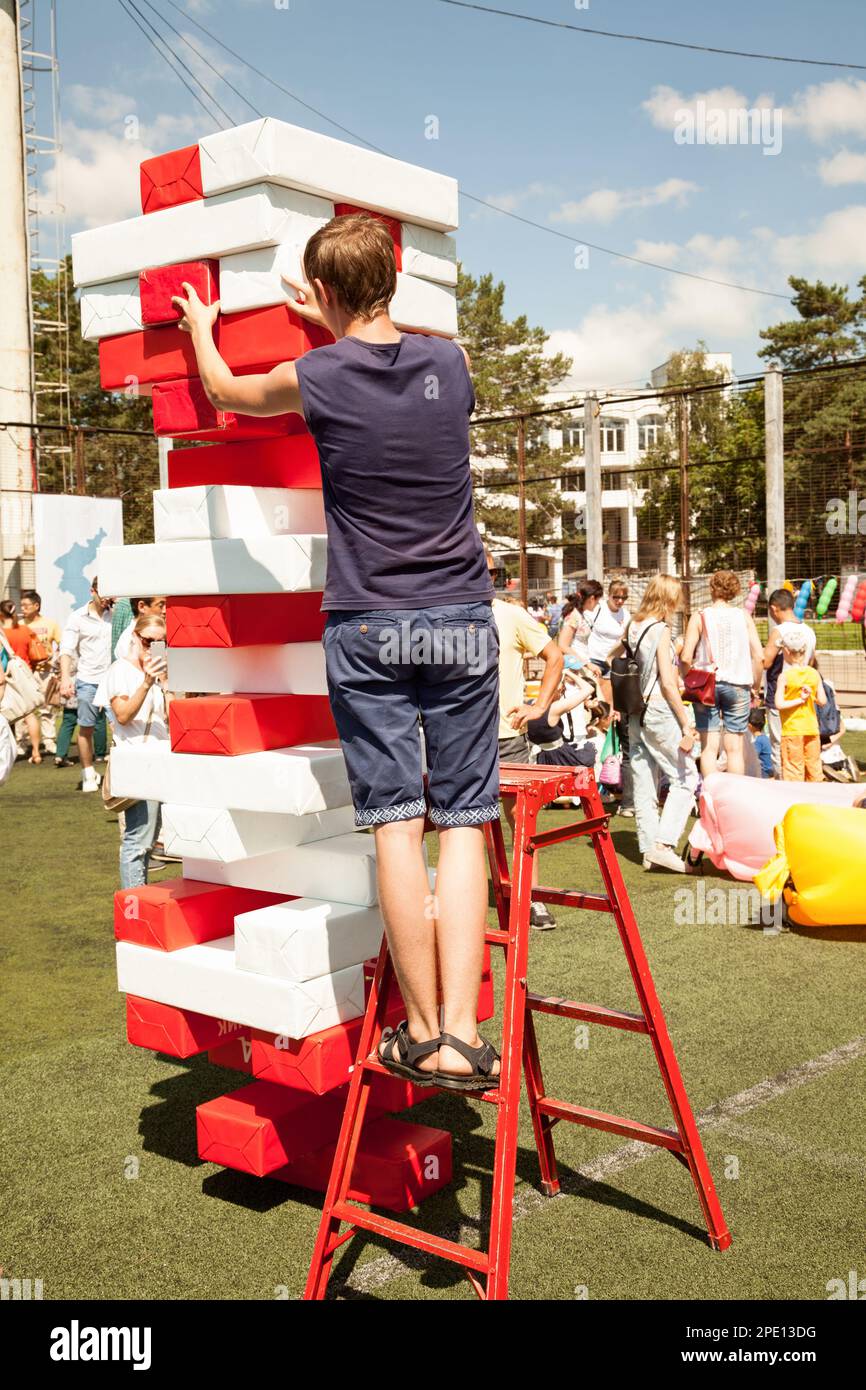 Khabarovsk, Russia - August 13, 2017: Young man play giant jenga game outdoors using a ladder. Person building a big tower of red and white bricks pop Banque D'Images