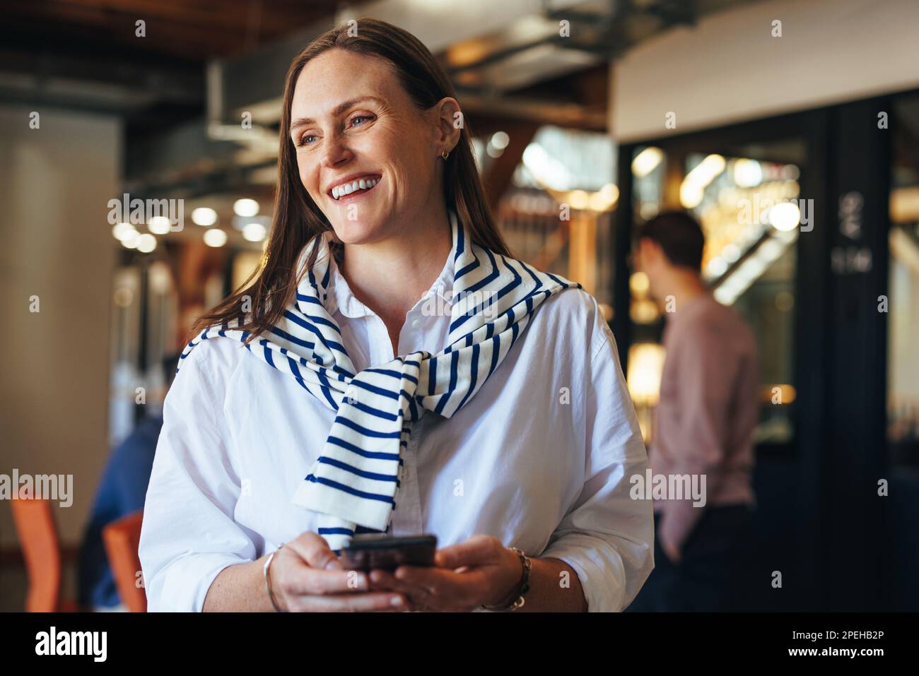 Femme d'affaires heureuse tenant un téléphone portable dans un lieu de travail. femme d'affaires debout dans un bureau avec ses collègues en arrière-plan. Banque D'Images