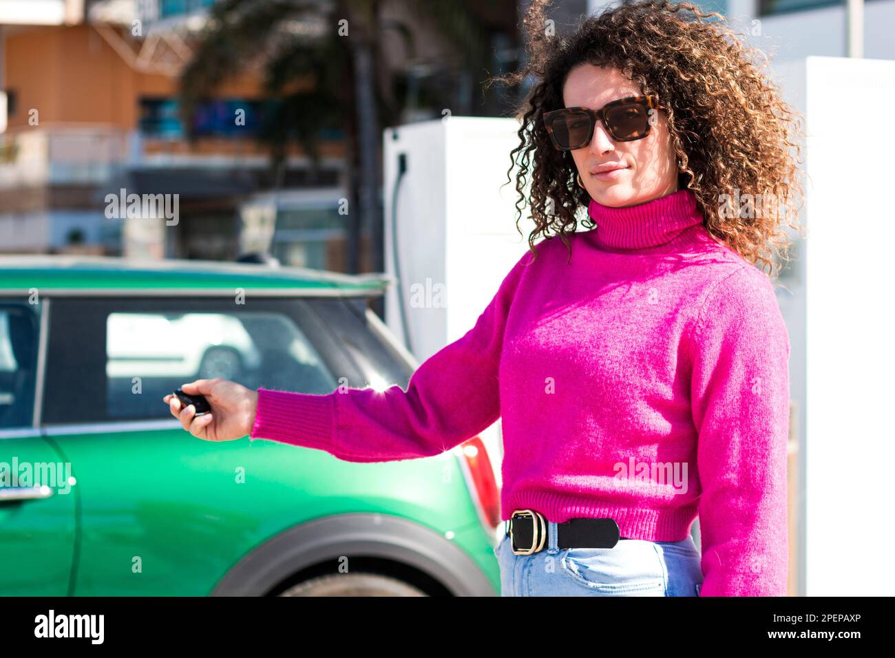 Jeune femme à poil dur positive en lunettes de soleil et tenue décontractée debout dans la rue près de la voiture verte en appuyant sur les touches d'envoi de signal lors de l'ouverture du véhicule Banque D'Images