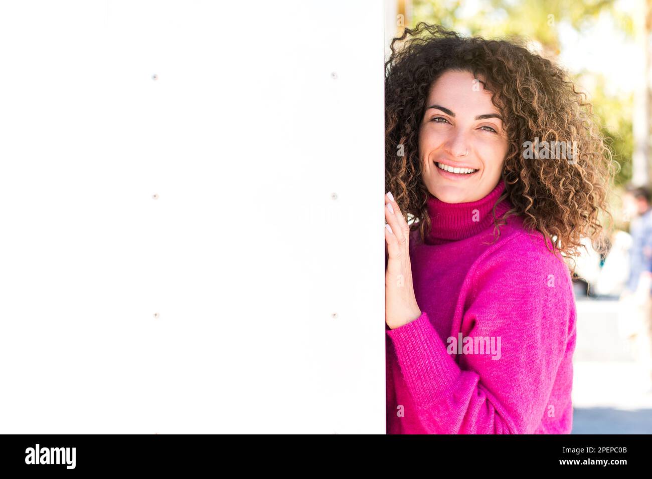 Heureuse femme aux cheveux bouclés portant un chandail à col roulé rose penché sur le mur tout en se tenant sur le fond flou de la rue de la ville le jour ensoleillé et lo Banque D'Images