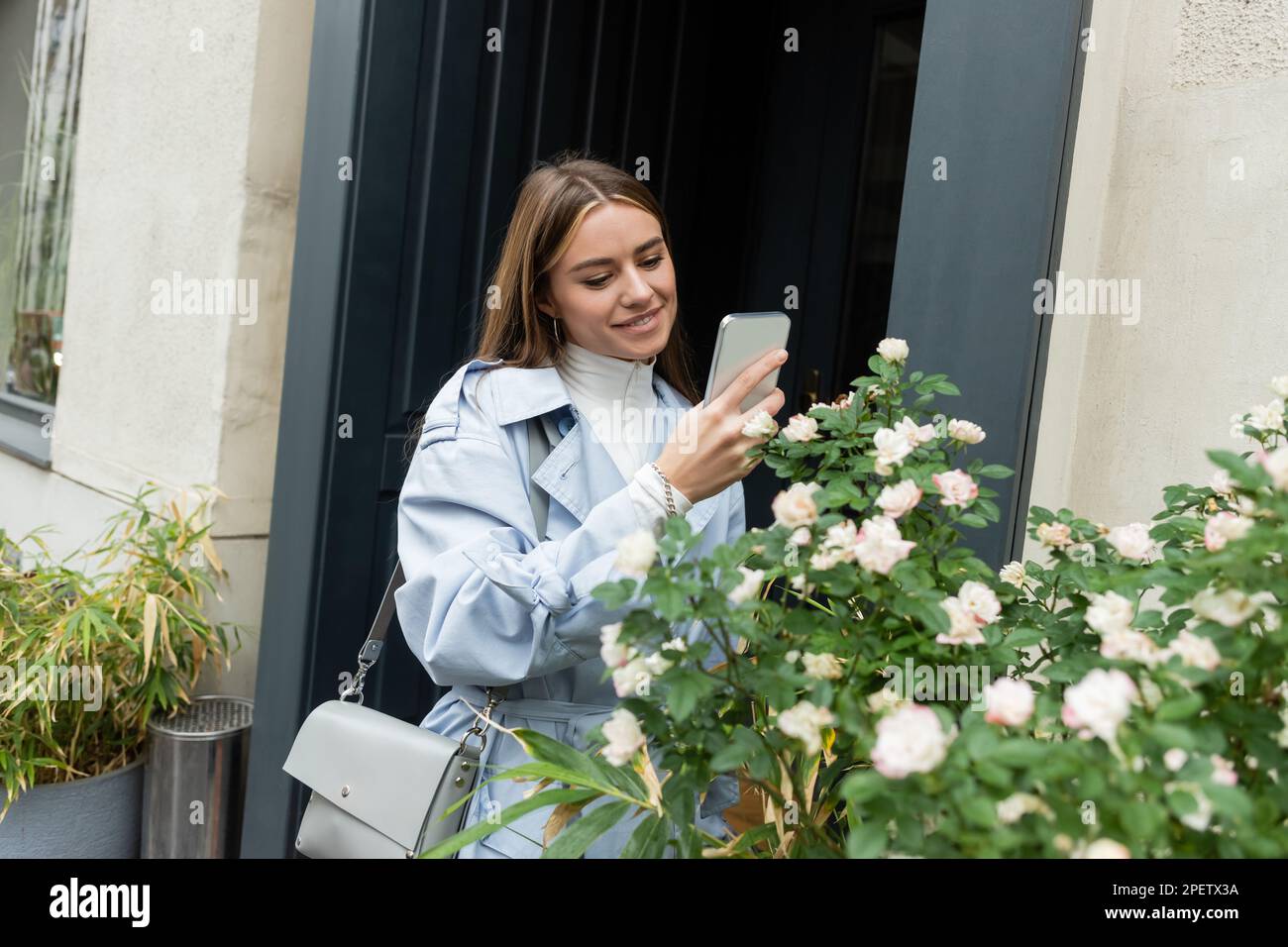 Bonne jeune femme en manteau de tranchée bleu prenant la photo du Bush vert  avec des fleurs fleuries dans la rue à Vienne Photo Stock - Alamy