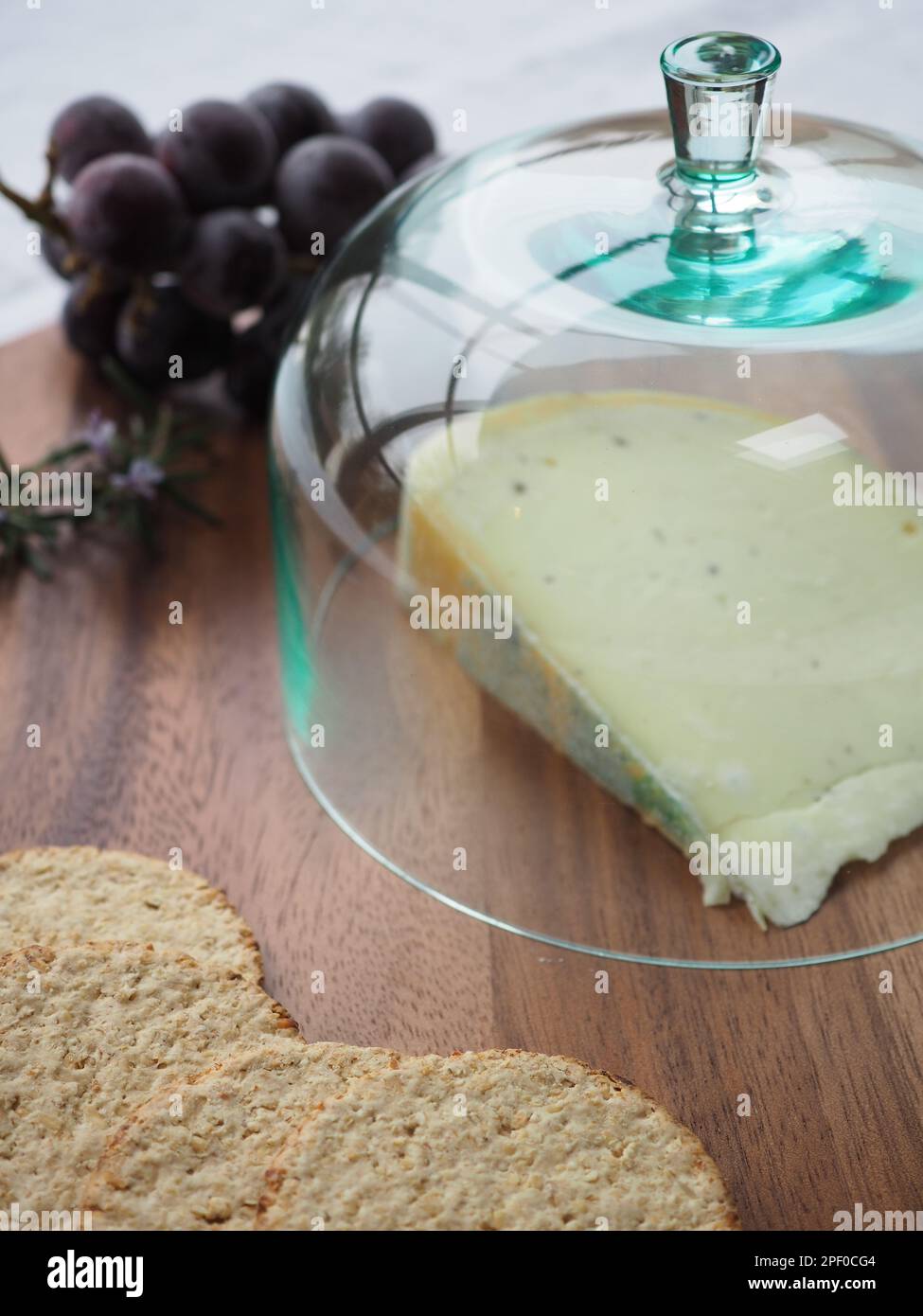 Gros plan d'un coin de fromage gouda sur une planche en bois sous un cloche ou un dôme en verre (cloche) avec des gâteaux d'avoine et des raisins violets sur le côté Banque D'Images