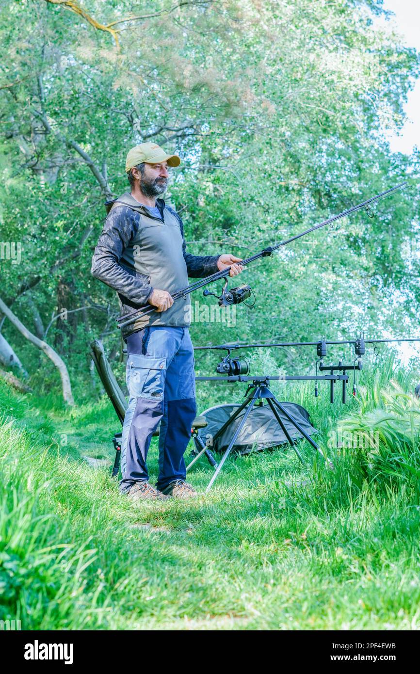 Pêcheur barbu mettant en place son équipement de pêche sur la rive de la rivière sur un champ d'herbe verte Banque D'Images