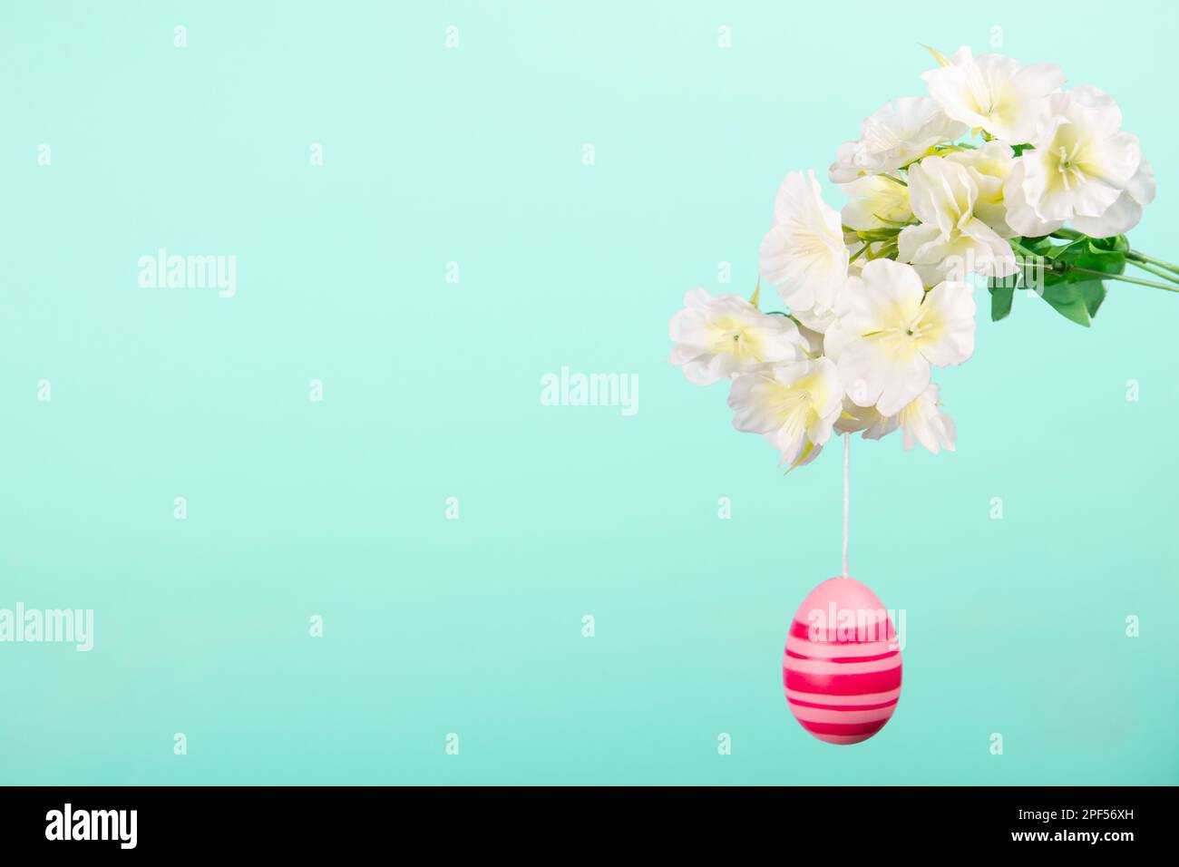 Un joli œuf de Pâques pastel accroché à une branche pleine de fleurs printanières sur fond turquoise pâle. Banque D'Images