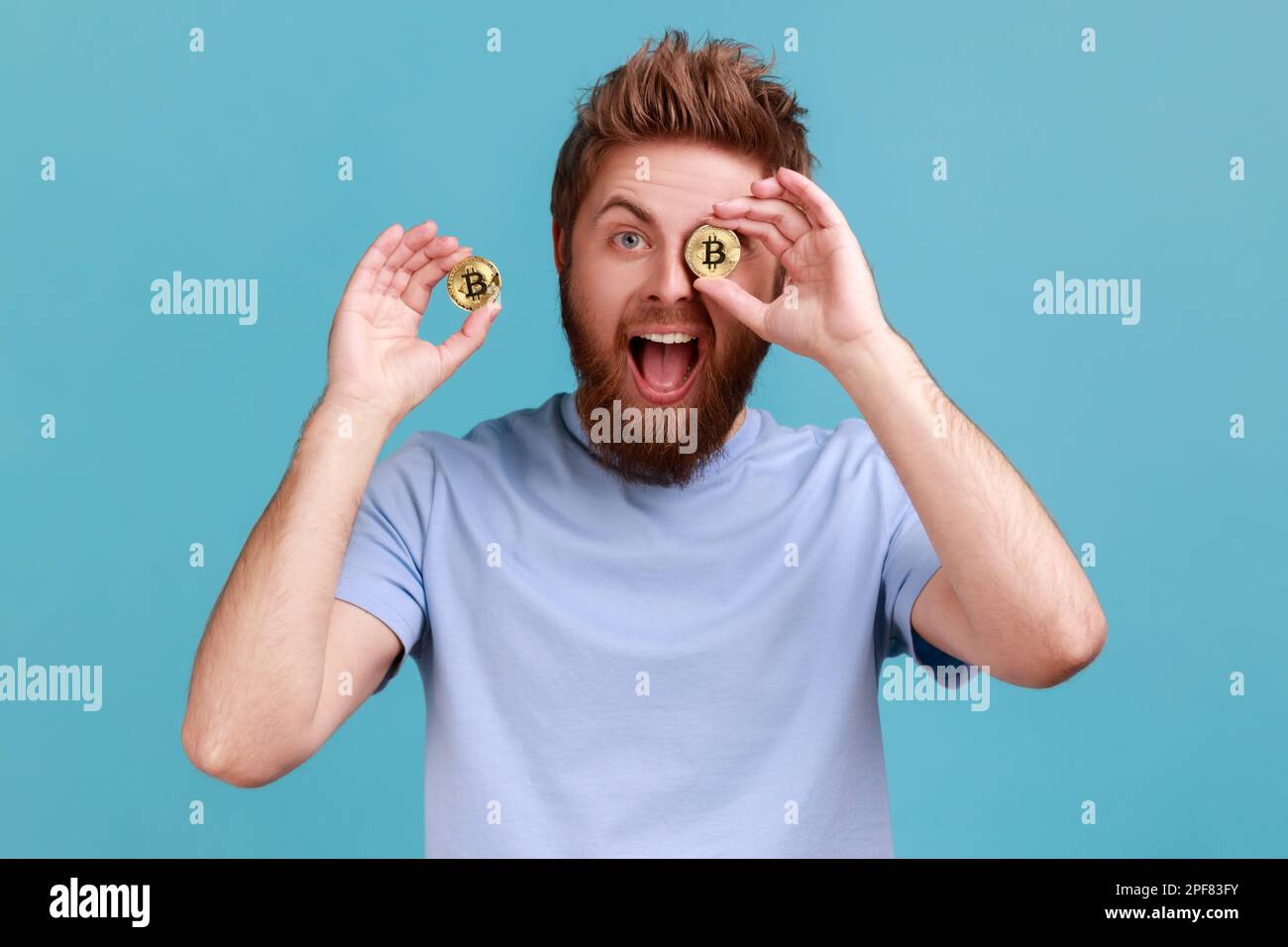 Portrait de l'homme barbu positif excité avec la bouche ouverte couvrant l'oeil avec la pièce de btc dorée et souriant à la caméra, publicité de crypto-monnaie. Studio d'intérieur isolé sur fond bleu. Banque D'Images