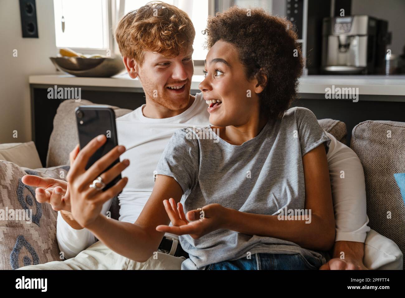 Jeune beau redhead souriant homme embrassant et regardant sa belle amie africaine heureuse avec téléphone, tandis qu'ils sont assis sur un canapé dans la vie confortable Banque D'Images
