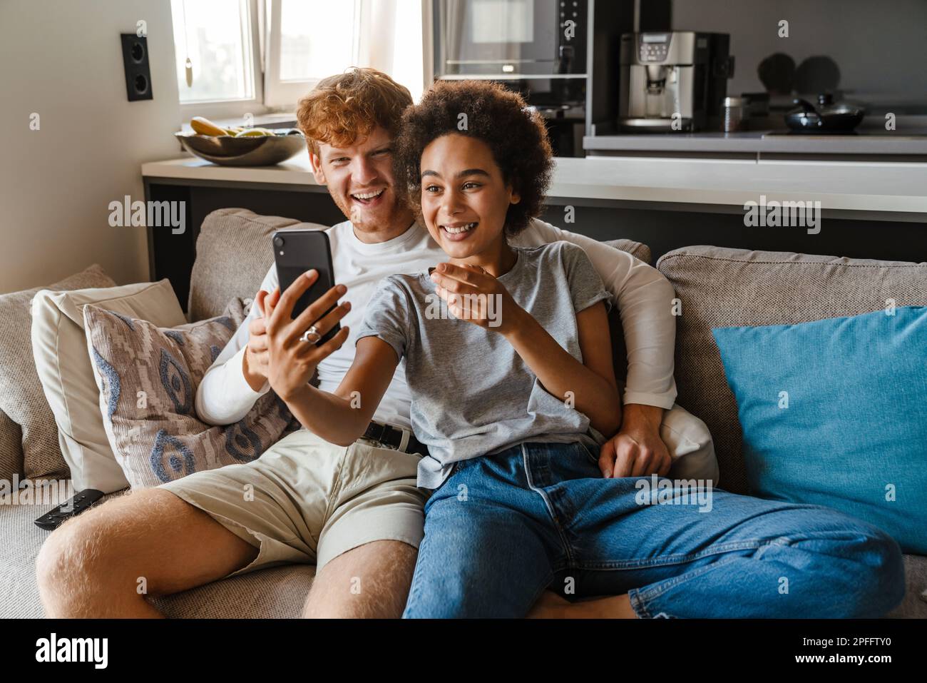 Jeune heureux couple interracial prenant selfie avec téléphone ensemble, tout en étant assis sur un canapé dans le salon confortable à la maison Banque D'Images
