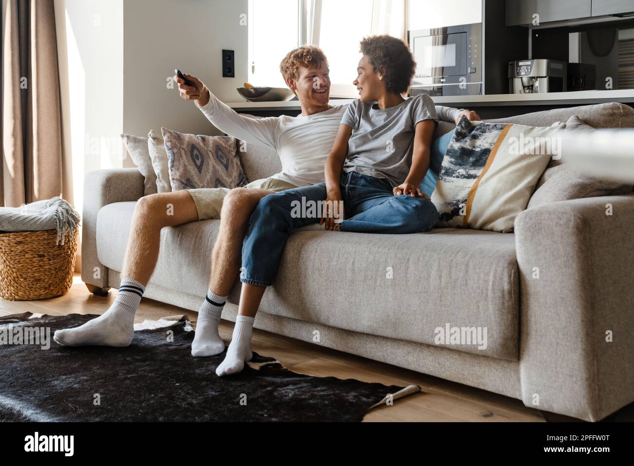 Jeune beau redhead souriant homme regardant sa belle petite amie africaine et changer de chaîne de télévision avec télécommande , pendant qu'ils sont assis toget Banque D'Images
