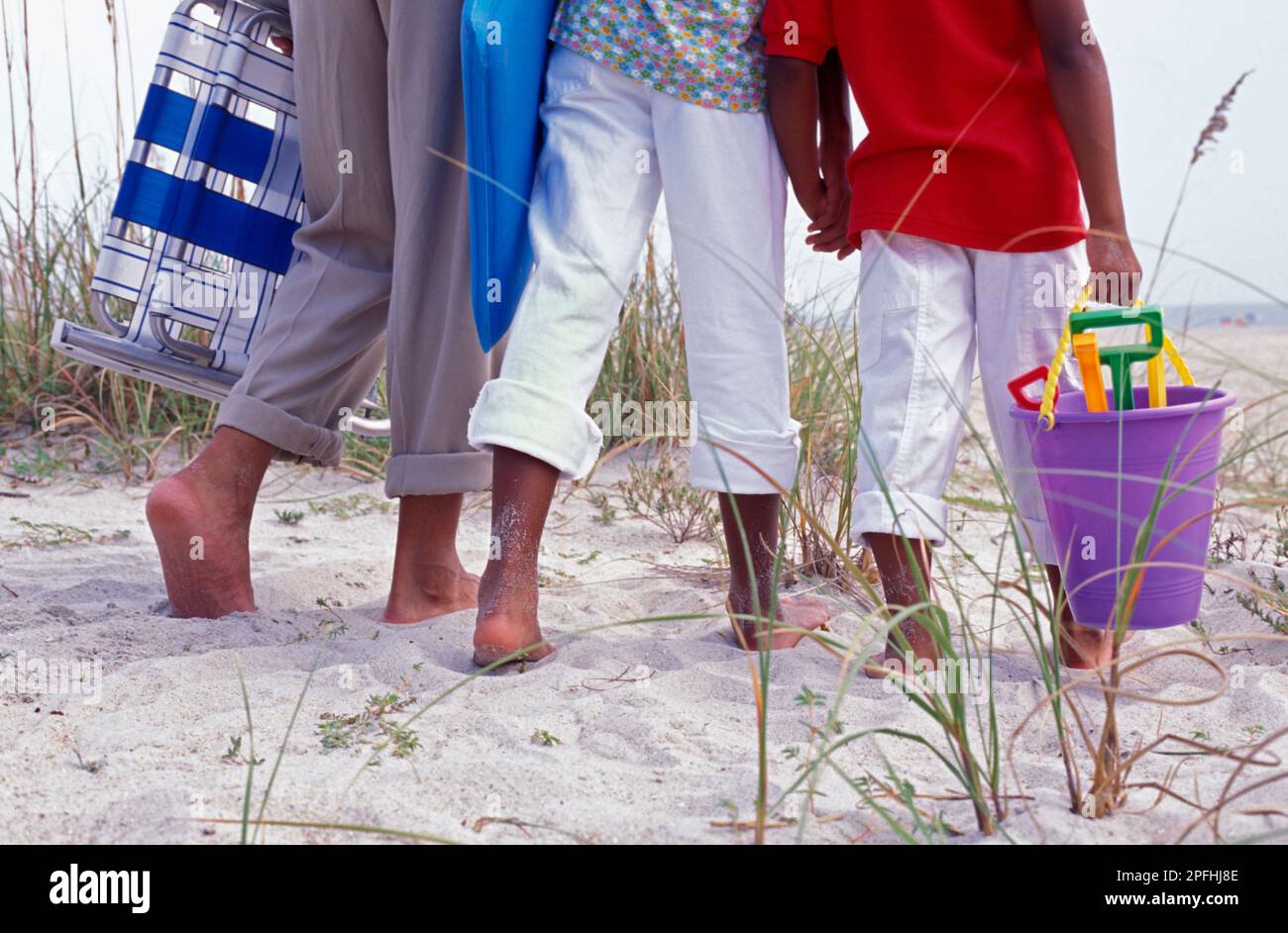 Une famille afro-américaine marchant sur une dune sur le chemin de la plage portant des chaises de plage et un seau Banque D'Images