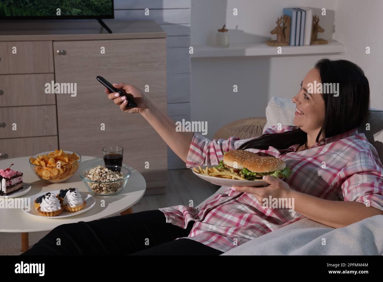 Bonne femme en surpoids avec de la nourriture malsaine regardant la télévision sur le canapé à la maison Banque D'Images