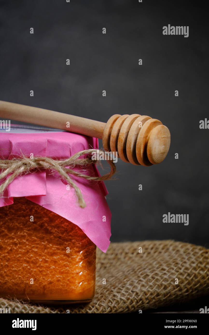 Un pot en verre de miel de fraise et une cuillère à miel se tiennent sur un toile de jute sur fond sombre Banque D'Images