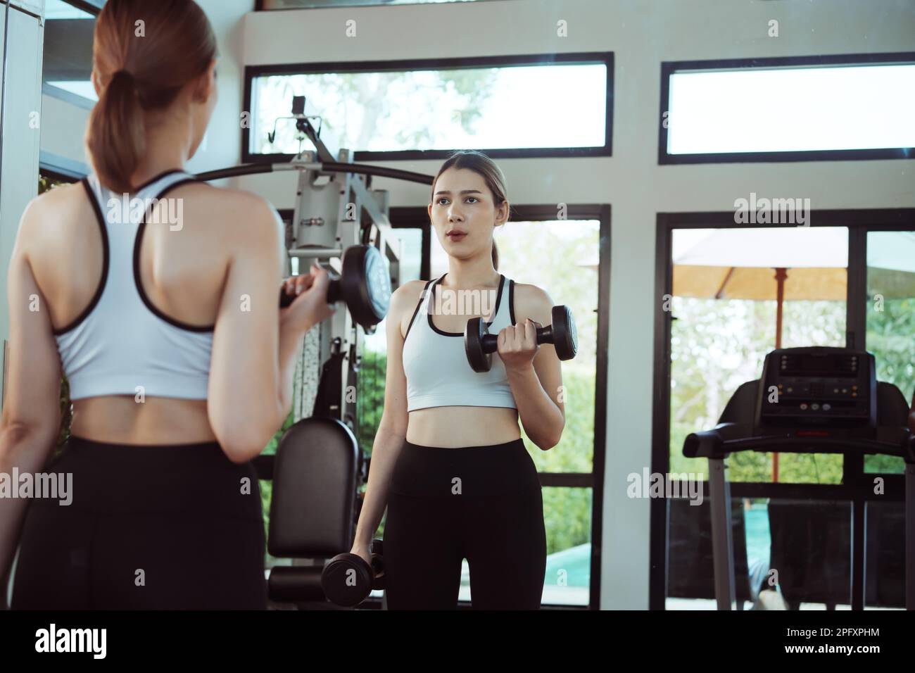 Jeune femme faisant de l'exercice avec haltère devant un miroir dans la salle de gym. Banque D'Images