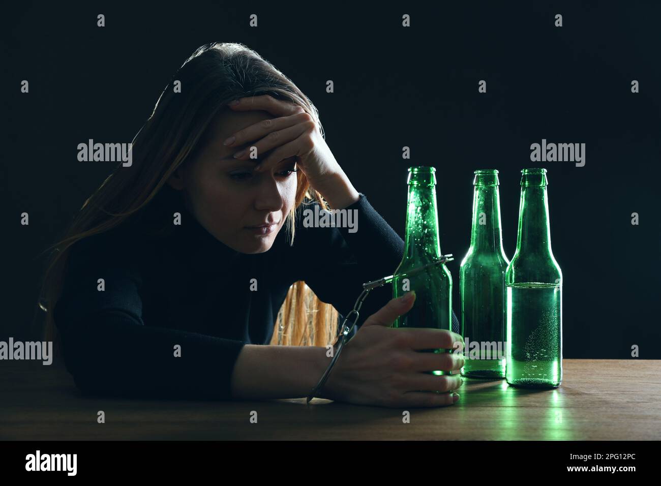 Dépendance à l'alcool. Femme menottée à une bouteille de bière à une table en bois sur fond noir Banque D'Images