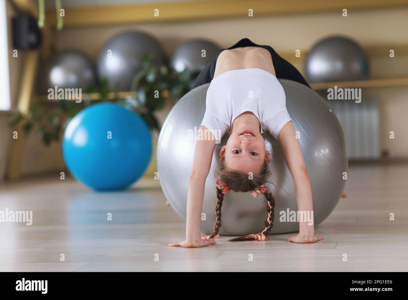 Une petite fille de vêtements de sport s'entraîne sur un fitball dans un studio de fitness, exécute un exercice de pont, l'enfant sourit et regarde la caméra Banque D'Images