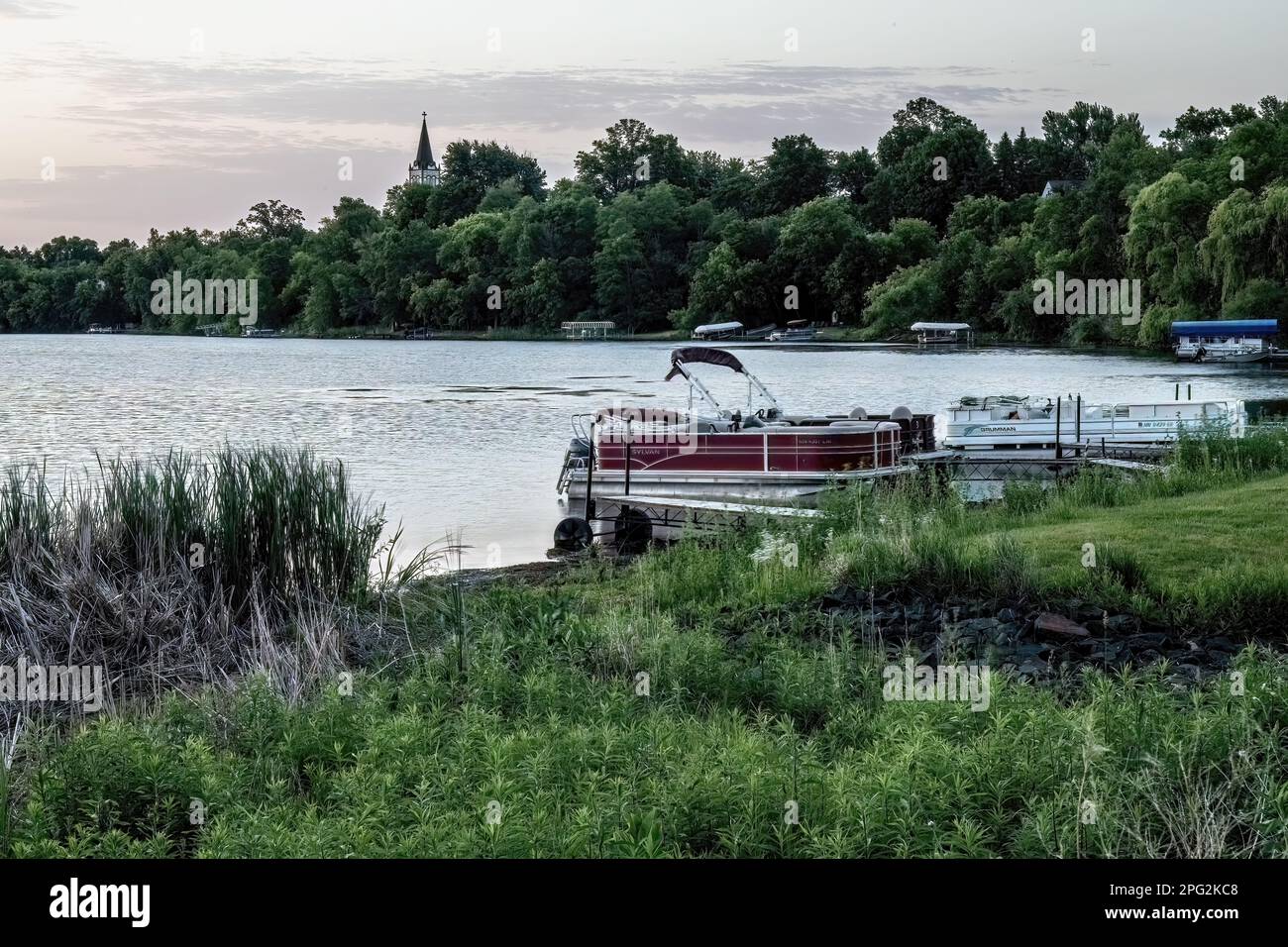 Pontons en bateau le long de la rive du lac North Center avec le clocher de l'église luthérienne du lac Chisago niché dans les arbres au-delà, lors d'une soirée de printemps. Banque D'Images